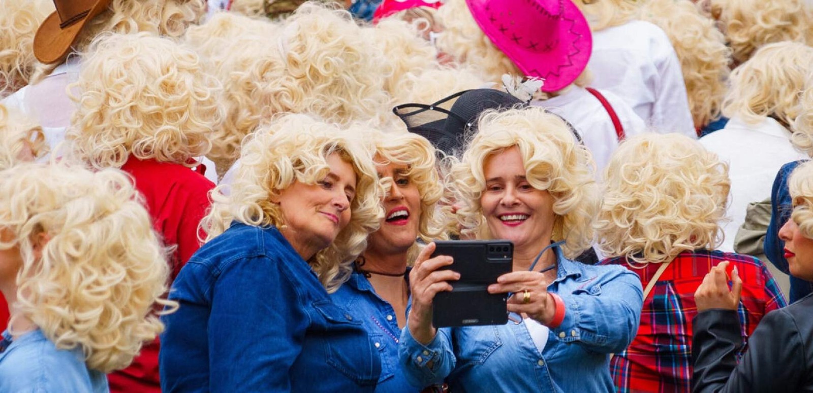 Three women in denim and blonde wigs taking a selfie in the middle of the crowd